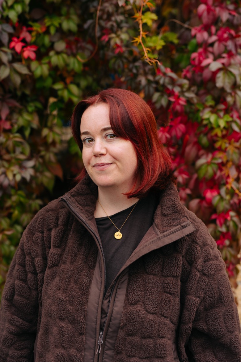 Jessica stands in front of a wall of red and green autumn leaves. She has shoulder-length red hair and is wearing a brown fleece jacket and a gold pendant necklace.