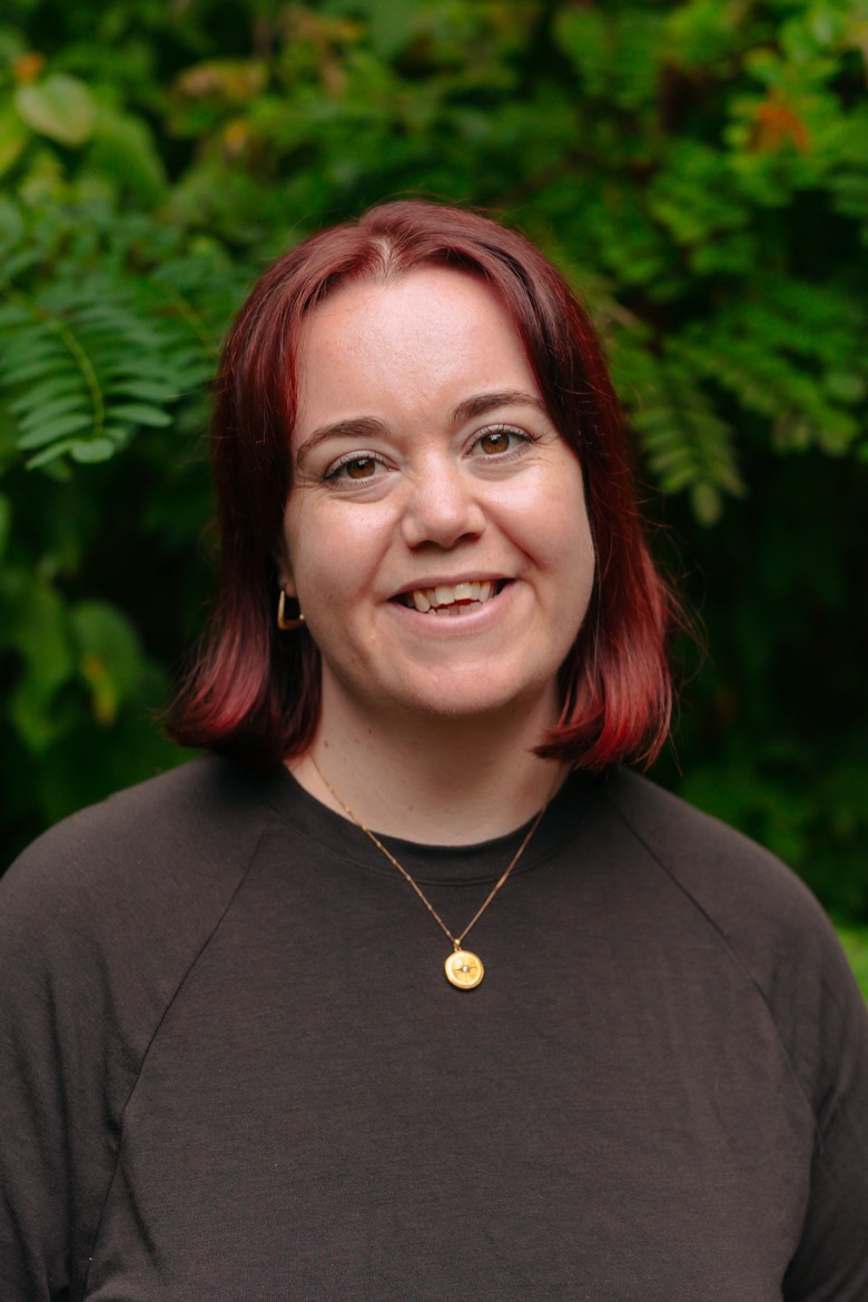 Jessica smiles in front of lush green foliage. She has shoulder-length red hair and is wearing a dark brown top and a gold pendant necklace.