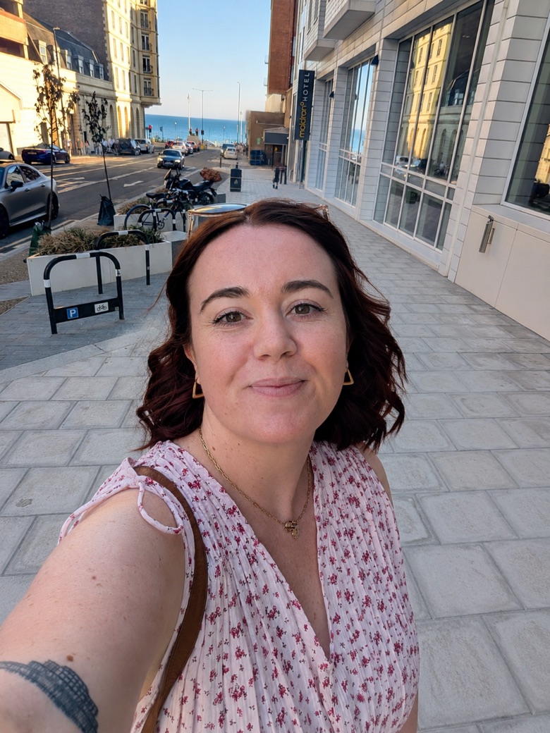 Jessica takes a selfie on a sunny street leading down to the sea. She has wavy auburn hair and is wearing a pink floral top.