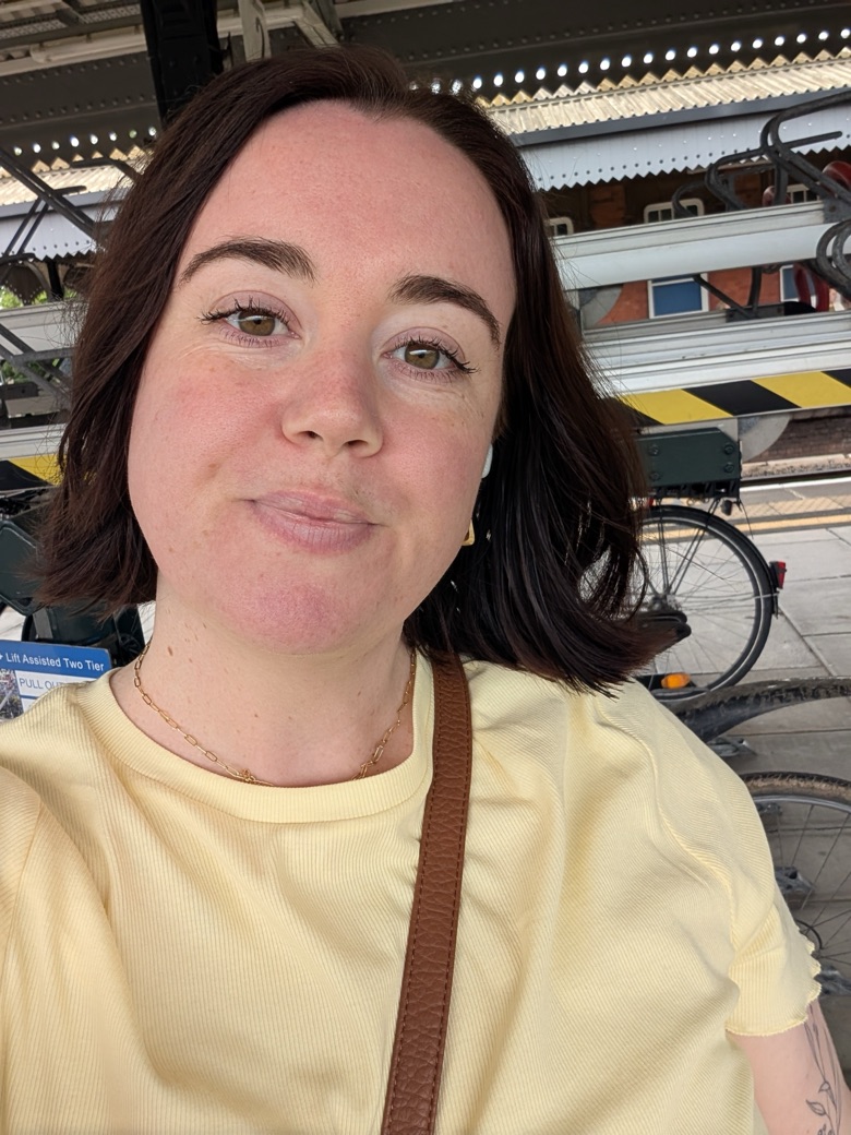 Jessica takes a selfie on a train station platform. She has shoulder-length dark hair and is wearing a pale yellow t-shirt and a gold necklace.
